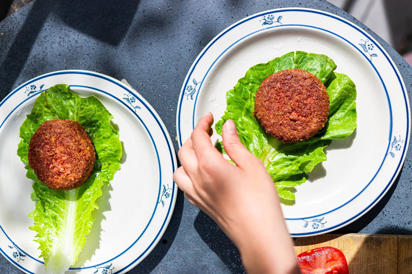 Cooked vegan meat sausage patty on plate with romaine lettuce leaf and tomatoes sliced for burger with female hand arranging two dishes